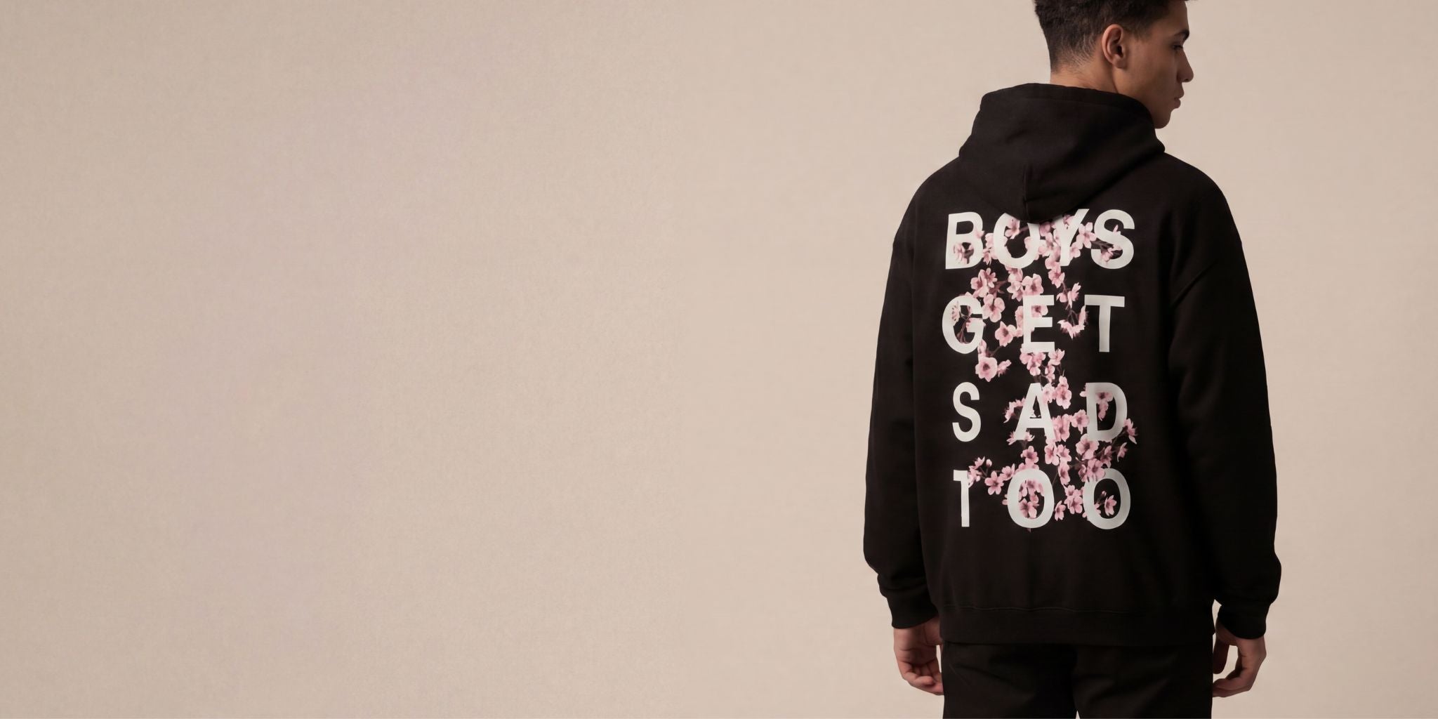 Model wearing a black hoodie viewed from the back, featuring large white text with pink floral graphics spelling ‘BOYS GET SAD TOO’ on the back against a neutral beige background.